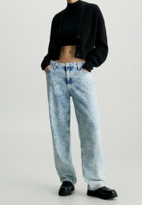 Černý zkrácený cardigan s ribbed texturou, spárovaný se světlými modrými volnými džíny z denimu s vybledlým a opotřebovaným vzhledem.