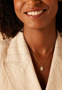 Gouden ketting met een ovale, doorzichtige stenen hanger versierd met kleine rhinestones, gedragen over een lichtgestreepte linnen blazer.