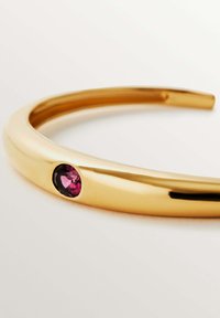 Brazalete de oro con forma redonda y suave que presenta una única gema ovalada de color rosa incrustada en el lateral. Superficie reflectante con un acabado pulido.