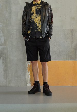 Giacca nera con zip e finitura opaca, camicia gialla e nera a fantasia, pantaloni corti neri e stivaletti neri con lacci e dettagli rossi.