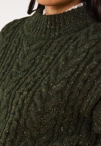 Maglione in filato a treccia verde scuro, con un collo alto e un motivo testurizzato con pezzetti di blu e oro sparsi su tutta la stoffa.