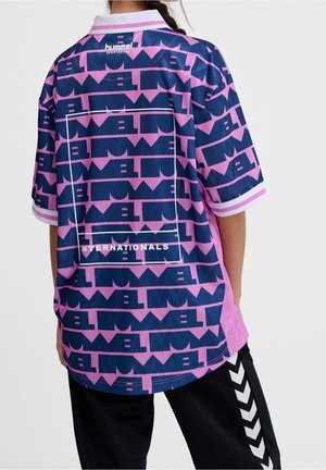 Osoba ubrana w różowo-granatową sportową koszulkę hummel z napisem "INTERNATIONALS", w połączeniu z czarnymi spodniami z białymi wzorami w kształcie szewronów.