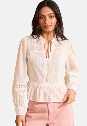 Blusa blanca de manga larga con peplum plisado, que presenta un tejido texturizado, detalles de encaje y un cuello abotonado con un lazo decorativo.