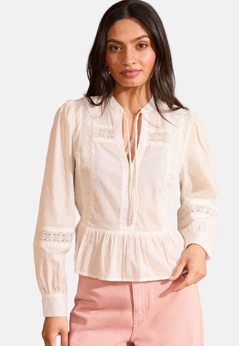 Chemisier blanc à manches longues avec un peplum plissé, présentant un tissu texturé, des accents en dentelle et une encolure boutonnée avec un détail de nœud.