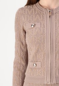 Lys brun strikket cardigan med teksturert mønster, med glidelås og to dekorative knapper på lommene.