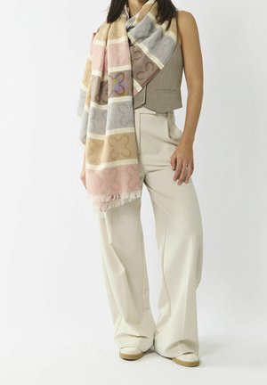 Femme portant un pantalon large beige, un gilet camel et une grande écharpe pastel à motifs floraux, debout devant un fond blanc.