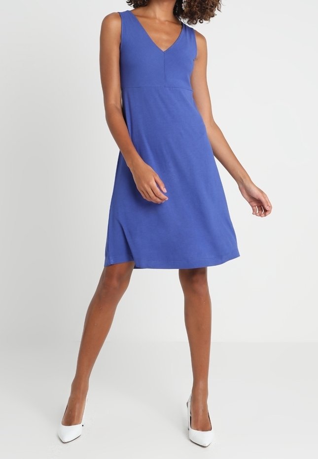 Femme portant une robe bleue sans manches, longueur genou, avec un col en V et des talons hauts blancs à bout pointu, debout sur un fond clair.