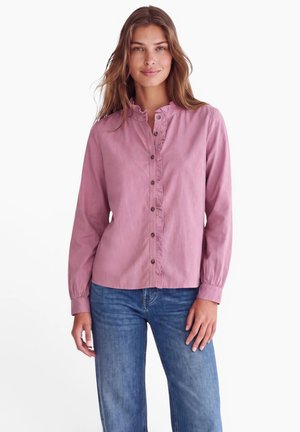 Kvinde med langt brunt hår iført en lyserød skjorte med rynker og blå jeans, stående mod en hvid baggrund.