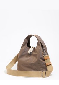 Bolsa tote de tela ligera marrón con correa ajustable beige y cierre de lazo de tela blanca sobre un fondo blanco.