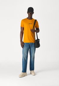 T-shirt di cotone gialla con piccolo logo, jeans blu a vestibilità rilassata, scarpe beige, una cuffia nera e una borsa nera, in piedi su uno sfondo neutro.