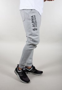 Graue Sweatpants mit engem Schnitt, die ein schwarzes Logo "ALPHA INDUSTRIES" an der Seite haben. Getragen mit schwarzen und grauen Sneakers.