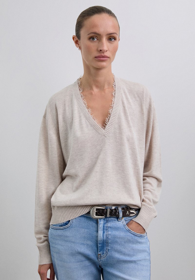 Suéter de punto beige con cuello en V y bordes deshilachados. Manga larga y dobladillo acanalado, combinado con jeans de mezclilla azul claro y un cinturón trenzado.