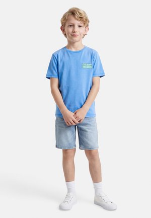 Młody chłopiec stoi z złączonymi dłońmi, ubrany w jasnoniebieską koszulkę, dżinsowe szorty, białe skarpetki i białe buty sportowe na białym tle.