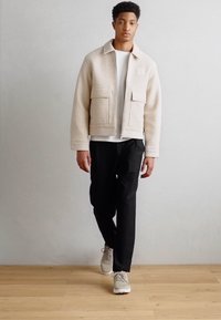 Beige színű fleece dzseki, két elülső zsebbel és cipzárral, fekete nadrággal és világosszürke sportcipőkkel párosítva, egy fa padlón.