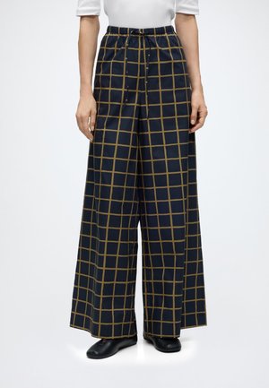 Pantalon taille large bleu marine avec motif quadrillé marron, taille élastique avec cordon, porté avec des chaussures noires et un haut blanc.
