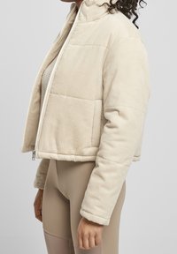 Beige kordjacka med en kort design, ribbad textur, ståkrage, framdragkedja och sidofickor, bärs över en ljus tröja.