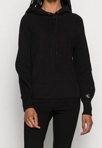 Czarna bluza z kapturem z teksturowanym wzorem na dzianinie, z prążkowanymi mankietami i dołem, regulowanymi sznurkami oraz małym haftowanym logo.