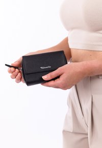 Moteris dėvi smėlio spalvos aprangą, laiko juodos odinės piniginės su užtrauktuku, ant kurios priekyje yra sidabrinis „Tamaris“ logotipas.