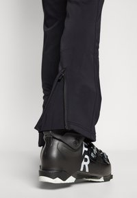 Sorte skibukser med glidelås ved leggen, laget av et glatt, teksturert materiale, kombinert med en svart skistøvel med hvite merker.