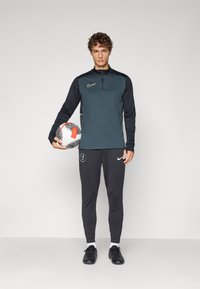 Nike donkerblauw langemouwen top met halve rits en gele accenten, gecombineerd met zwarte taps toelopende broek en zwarte voetbalschoenen, terwijl hij een gestreepte voetbal vasthoudt.