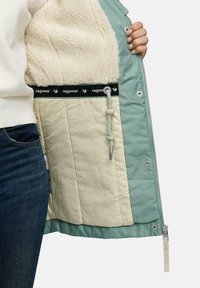 Zielona kurtka z pluszowym beżowym wykończeniem. Cechy obejmują zamek błyskawiczny, regulowany ściągacz oraz markową taśmę wzdłuż wewnętrznej szwu.