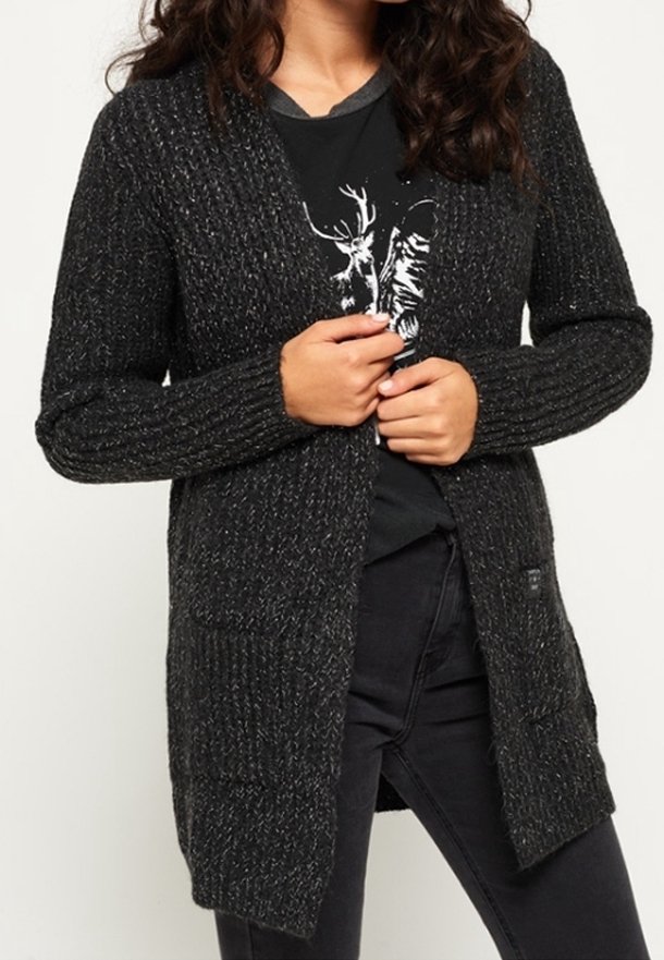Femme portant un cardigan en tricot gris foncé par-dessus un t-shirt noir à motif et un pantalon noir, les mains tenant le cardigan fermé.