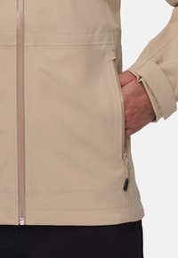 Hell-beige wasserdichte Jacke mit einem Frontreißverschluss, Seitentaschen und verstellbaren Manschetten. Glatte Textur und minimalistisches Design.