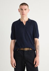 Marineblå polo med korte ærmer, blødt materiale, kravedesign, parret med mørke jeans og et lysebrunt bælte. Subtil branding synlig.