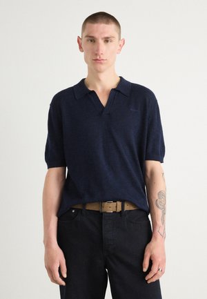 Navy polo shirt with short sleeves, soft texture, collar design, paired with dark jeans and a light brown belt. Subtle branding visible.