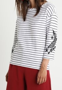 Wit gestreept shirt met driekwartmouwen, voorzien van zwarte bloemenborduursels op de mouwen. Gecombineerd met rode wijde broek.