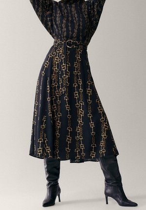 Une longue robe noire avec un motif en chaîne doré, dotée d'une ceinture cintrée à la taille et d'une jupe fluide, associée à des bottes noires montant jusqu'aux cuisses.