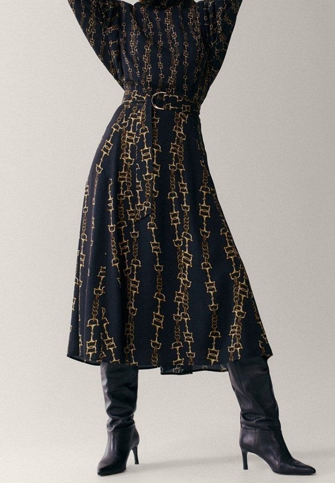 Une longue robe noire avec un motif en chaîne doré, dotée d'une ceinture cintrée à la taille et d'une jupe fluide, associée à des bottes noires montant jusqu'aux cuisses.
