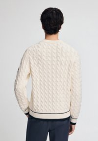 Kabelstrickpullover in Off-White, mit Rundhalsausschnitt, gerippten Ärmelbündchen und Saum mit marineblauen Streifen, der ein strukturiertes Design zeigt.