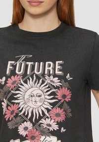 Zwarte katoenen t-shirt met een grafisch ontwerp van een zon, bloemmotieven in roze en wit, en de tekst "De TOEKOMST" erboven.