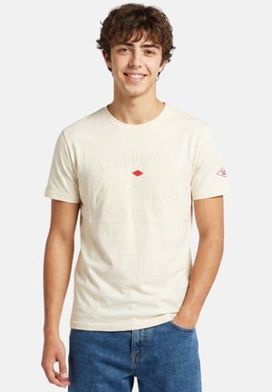 Jeune homme aux cheveux bruns bouclés, portant un t-shirt graphique couleur crème et un jean bleu, souriant avec une main dans la poche.