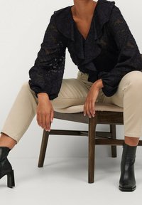 Schwarze Spitzenbluse mit Rüschenausschnitt und puffigen Ärmeln, kombiniert mit beigen, schmal geschnittenen Hosen. Das Modell trägt schwarze, knöchelhohe, hohe Stiefel.