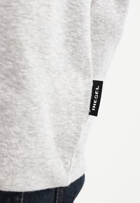 Sudadera gris con una textura suave y una etiqueta negra con texto blanco en el dobladillo. El diseño es simple con un corte holgado.