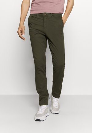 Chinos vert olive avec une coupe slim, dotés d'une fermeture à bouton à l'avant et de deux poches latérales. Portés avec des baskets blanches et une chemise rose clair.