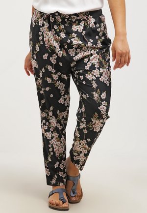 Czarne spodnie zwężane z jasnoróżowym kwiatowym nadrukiem noszone z białą bluzką i niebieskimi sandałami z odkrytymi palcami na jasnym tle.