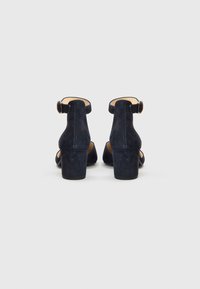 Tumši zili zamša apavi ar papēža siksniņu, aizvērtu aizmuguri un kuplu papēdi. Gluda tekstūra ar apaļā purngala dizainu un metāla sprādzes detaļām.