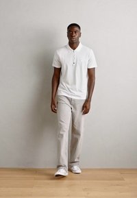 Hvid kortærmet polo shirt med knaplukning og patch-logo, parret med lys grå lige ben bukser og hvide sneakers.