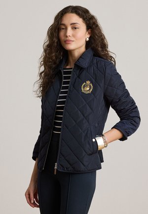 Žena kučeravých vlasov, ktorá má na sebe námornícku prešívanú bundu s erbom, pruhované tričko, čierne nohavice a strieborné náramky na ľavom zápästí.