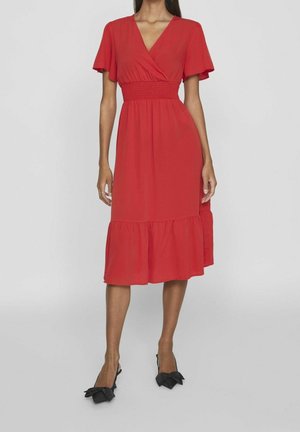 Vestito rosso avvolgente con maniche a sbuffo corte, vita arricciata e orlo svasato. Indossato con scarpe slingback nere con dettagli a fiocco.