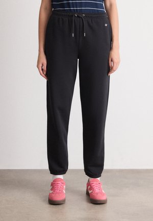 Person wearing black drawstring sweatpants and pink sneakers standing on a gray floor against a white wall.