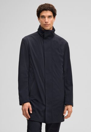 Homme aux cheveux bouclés portant un manteau noir mi-long à col montant, debout devant un fond gris clair uni.