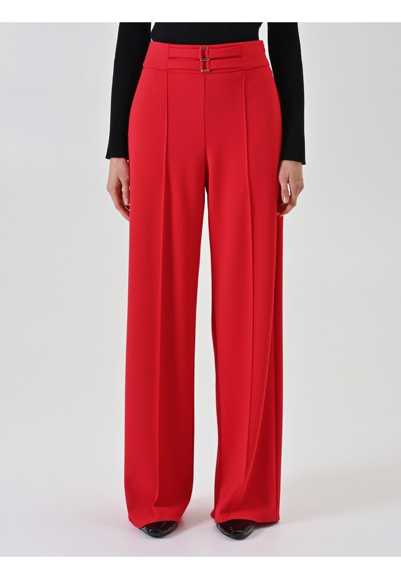 Frau in rot gestreiften, hoch taillierten, weiten Hosen mit doppeltem Schnallenbund und schwarzen spitzen Schuhen, stehend vor einem einfarbigen Hintergrund.