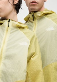 Deux personnes portant des vestes légères à capuche jaunes avec le logo The North Face, visibles de la poitrine jusqu'au menton sur un fond blanc.