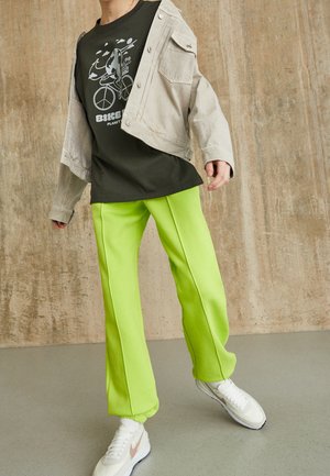 Personne portant un pantalon vert néon, un t-shirt noir avec un motif de vélo, une veste beige et des baskets blanches, debout à l'intérieur sur un sol gris.