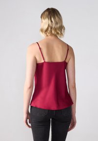 Camisola in satin bordeaux con spalline sottili, scollatura dritta e orlo curvato, abbinata a jeans scuri e attillati. Il tessuto testurizzato riflette la luce.