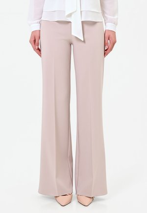 Donna che indossa pantaloni beige chiaro a gamba larga, camicetta bianca con dettaglio a fiocco e tacchi a punta color nude, in piedi su sfondo bianco.